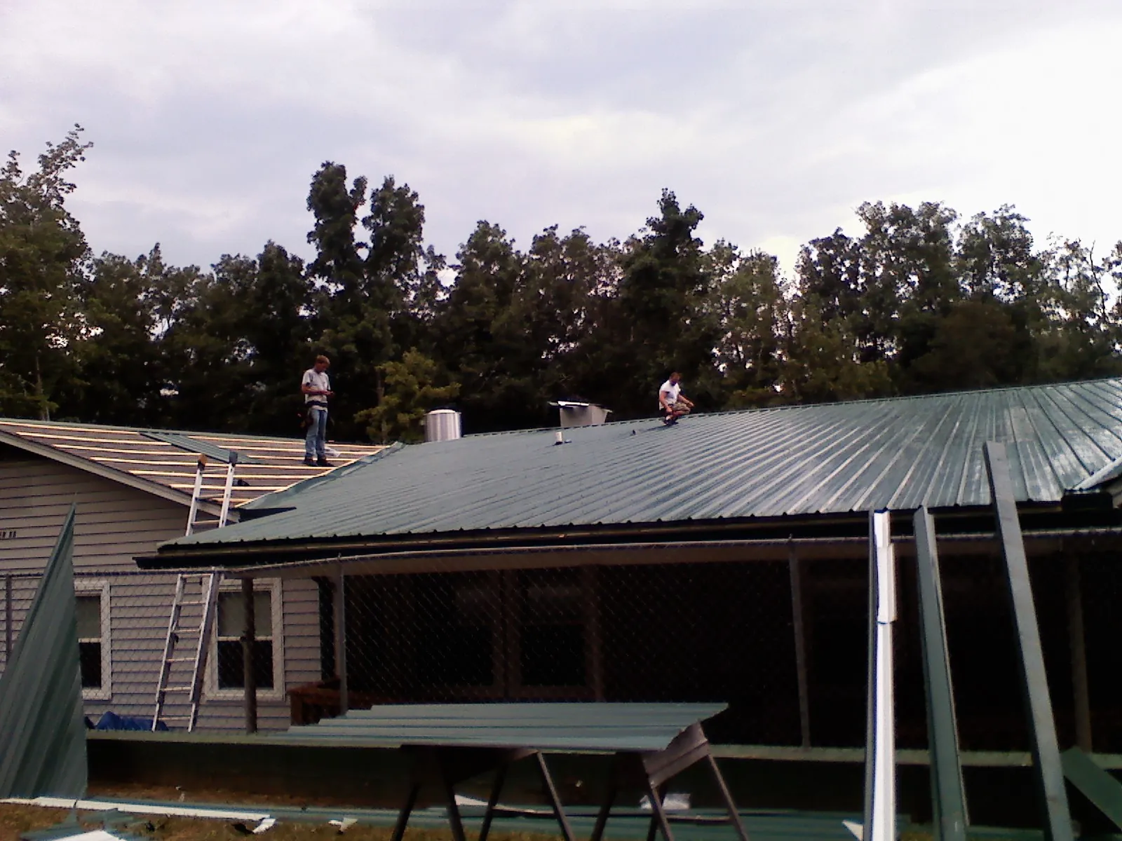 Two roofers installing green metal panels above a covered porch in Anderson